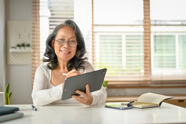 집 작업 공간에서 디지털 태블릿을 사용하여 웃고 행복한 60대 아시아계 여성