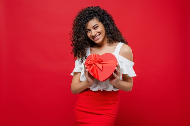 Smiling happy african woman holding heart shaped gift isolated on red