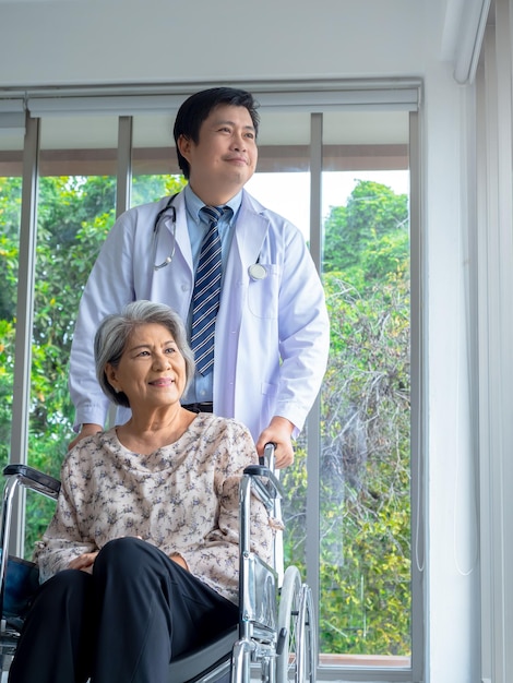 Smiling happy asian elderly senior female patient sit in\
wheelchair with man doctor in white coat standing in medical office\
in hospital vertical style caregiving healthcare and medical\
concept