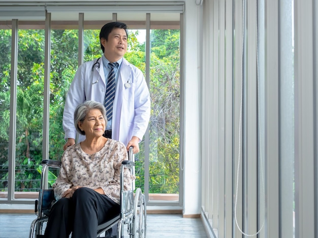 Smiling happy asian elderly senior female patient sits in\
wheelchair while man doctor in white coat standing in medical\
office in hospital vertical style caregiving healthcare and medical\
concept