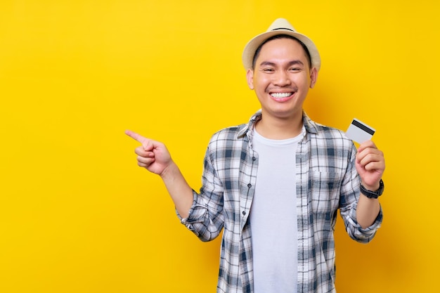 Smiling happy Asian ethnicity young man 20s wearing casual clothes hat holding in hand credit bank card pointing finger aside isolated on yellow background studio portrait People lifestyle concept