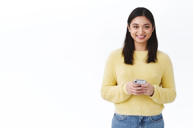 Smiling happy asian girl holding mobile phone and looking camera over white background, calling friend