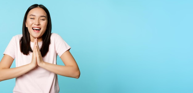 Photo smiling happy asian woman shows namaste beg gesture and dreaming of something making wish high hopes