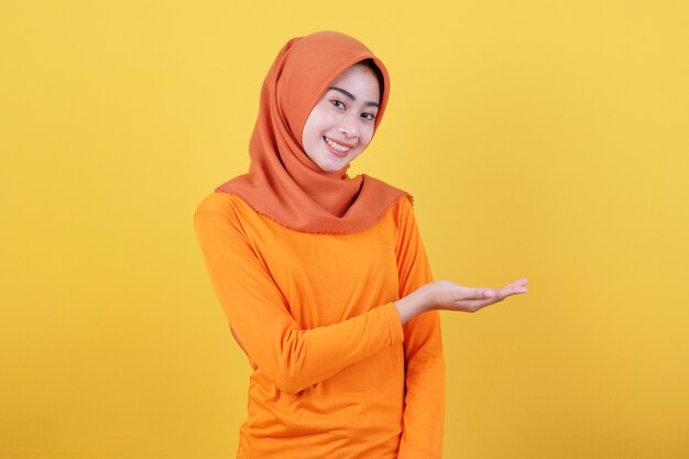 Smiling happy asian woman something in her hand and presenting it on light yellow banner background wearing hijab