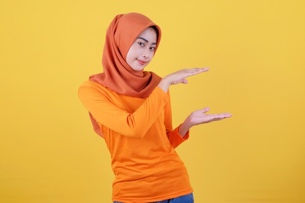 Smiling happy asian woman something in her hand and presenting it on light yellow banner background wearing hijab