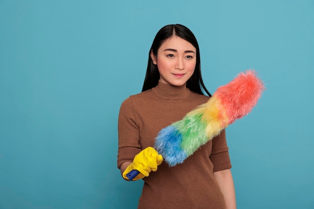 黄色のゴム手袋をはめた笑顔の幸せなアジアの若い女性が、青の背景にダストブラシを持ち、家の掃除のコンセプトを持ち、日々の雑用から陽気な楽観的で喜んでいる主婦