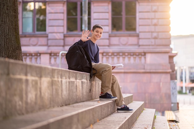 Улыбающийся счастливый мальчик с книгой в руке сидит на бетонных ступеньках и машет кому-то