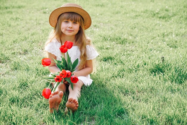 Улыбающаяся счастливая детская девочка с длинными светлыми золотыми волнистыми волосами в шляпе сидит на зеленой траве лужайки, держа в руках букет красных тюльпанов. Фото высокого качества