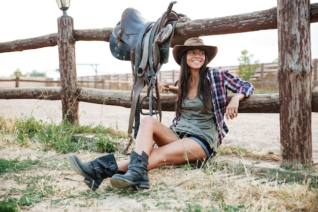 Photo smiling happy cowgirl in hat sitting and resting at the ranch fence