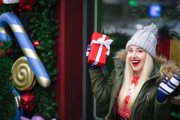 Smiling happy and excited blonde girl in a warm hat holding a gift box smiling and looking