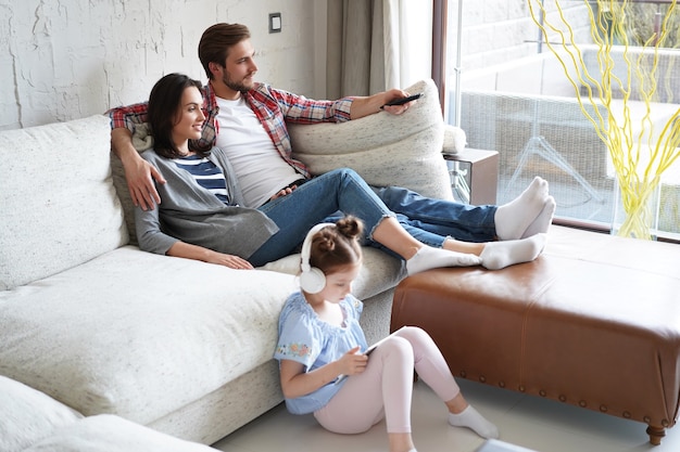 Smiling happy family sit relax on couch in living room watch little daughter drawing in digital tablet. Happy weekends at home.