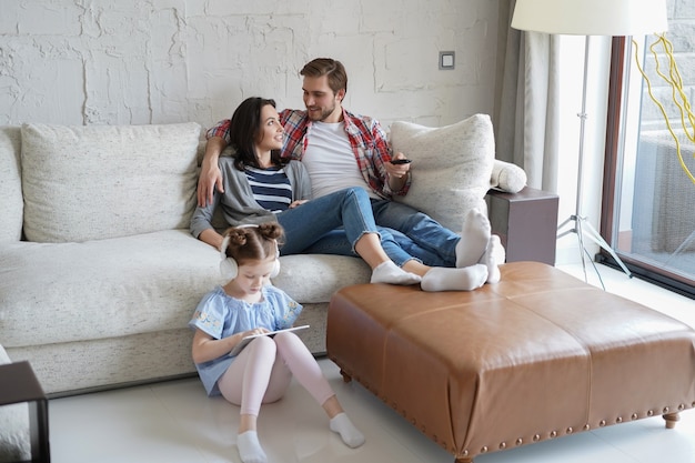 Smiling happy family sit relax on couch in living room watch little daughter drawing in digital tablet. Happy weekends at home.