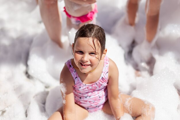 ピンクの水着を着た笑顔の幸せな女の子が、夏のホテルの泡パーティーでプールのそばに座っています...