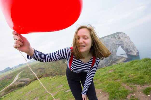 Smiling happy girl with a red balloon in the shape of a heart at background of scenery Etretat. France