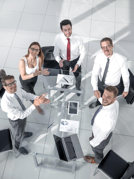 カメラを見上げてオフィスのテーブルの周りに座ってチームとして働く若いビジネスマンの笑顔の幸せなグループ