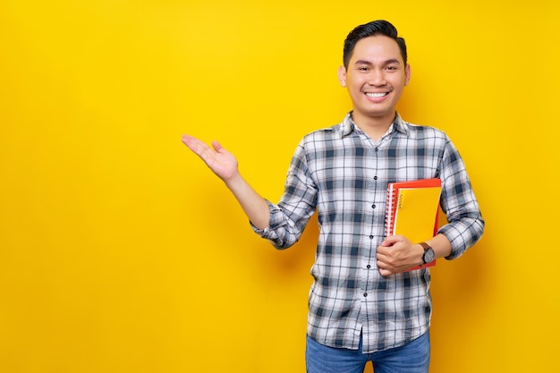 Smiling happy handsome young Asian man wearing a white checkered shirt holding books on hand and doing presentation isolated over yellow background People Lifestyle Concept