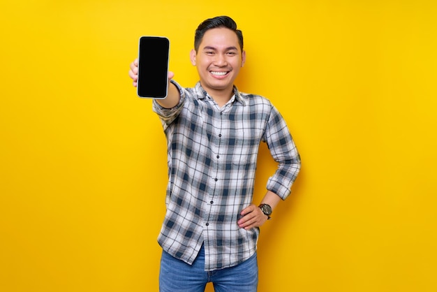 Smiling happy handsome young Asian man wearing a white checkered shirt showing mobile phone with blank screen for an advertisement isolated over yellow background People Lifestyle Concept