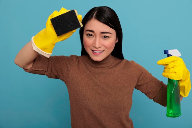 Smiling happy and joyful asian young woman holding a sponge with yellow gloves isolated on a blue background, Housewife worker, Cleaning home concept, Delighted laughing and positive state of mind