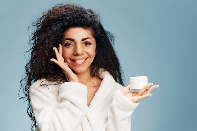 Smiling happy laughing curly latin lady in bathrobe looking at\
camera hold jar of cream touching cheek posing isolated on pastel\
blue background organic cosmetic product ad concept natural\
beauty