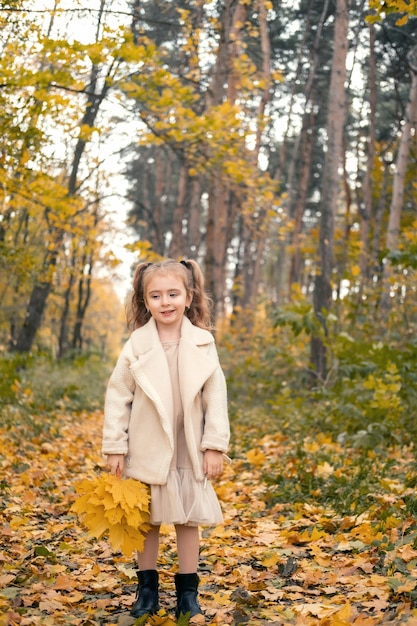 웃고 있는 행복한 어린 소녀 코트와 단풍을 들고 있는 드레스, 가을 숲에서 즐거운 시간
