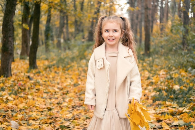 smiling happy little child girl in coat and dress holding autumn leaves, having fun in fall forest