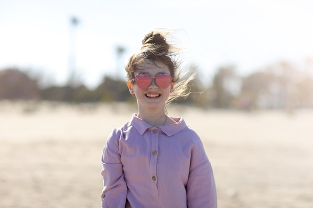 Фото Улыбающаяся счастливая маленькая девочка в солнечных очках в форме сердца веселится на пляже летние каникулы ребенок смотрит в камеру на фоне пальм