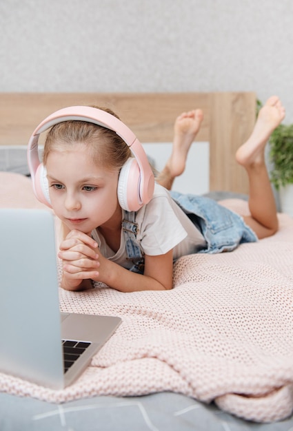 Smiling happy little kid girl using laptop in wireless headphones in bed at home.