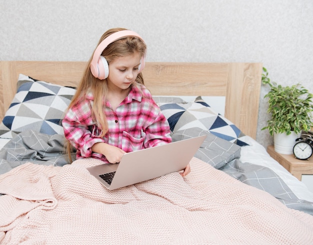 Smiling happy little kid girl using laptop in wireless headphones in bed at home