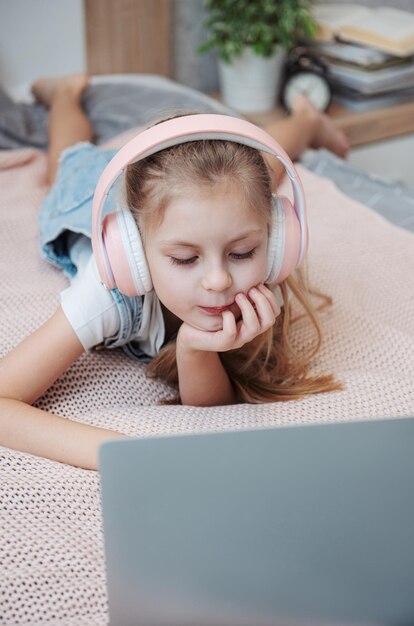 Smiling happy little kid girl using laptop in wireless headphones in bed at home