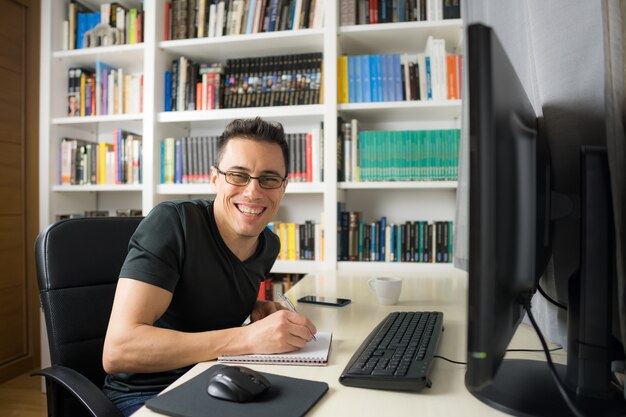 Smiling and happy man in his room sitting at the computer looking at camera, wearing a shirt. Mid shot.