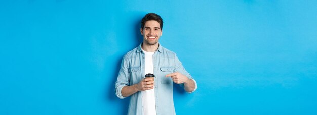 Smiling happy man pointing at paper cup with coffee recommending cafe standing over blue background