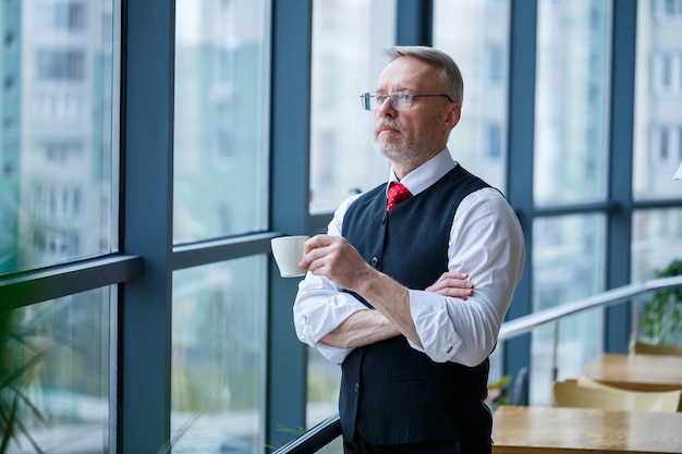 Smiling happy managing director thinks about his successful\
career development while standing with a cup of coffee in his hand\
in his office near the background of a window with copy space