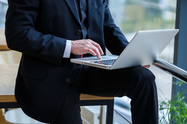 Photo smiling happy managing director thinks about his successful career development while standing with a laptop in his office near the background of a window with copy space