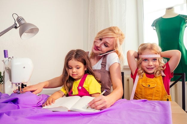 Smiling happy multitasking blonde mother teaches a little sisters to sew on a sewing machine by herself and making selfie pictures photos on smartphone