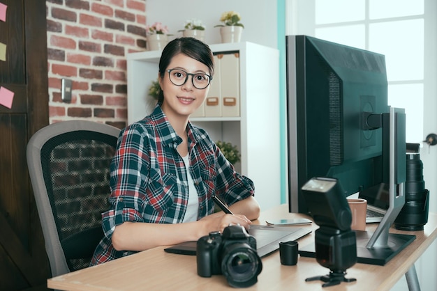 웃고 있는 행복한 사진가 여성은 디지털 펜 리터치 사진을 사용하여 작업용 책상에 앉아 있고 편집실에서 카메라를 마주하고 있습니다.