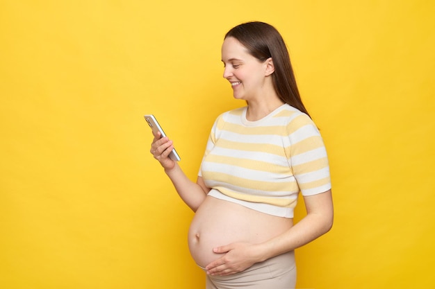 Smiling happy positive young Caucasian pregnant woman dressed in top posing against bright yellow wall holding smartphone browsing internet web pages