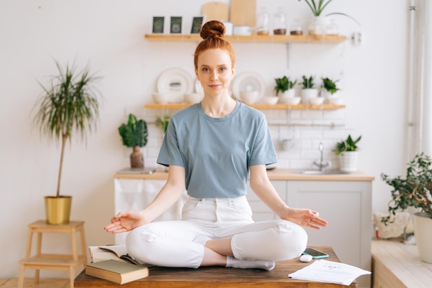 Smiling happy redhead young woman is meditating while sitting on desk at home office looking at camera Peaceful female sitting in lotus pose