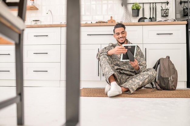 Smiling happy serviceman seated on the kitchen floor looking at the photo in the frame