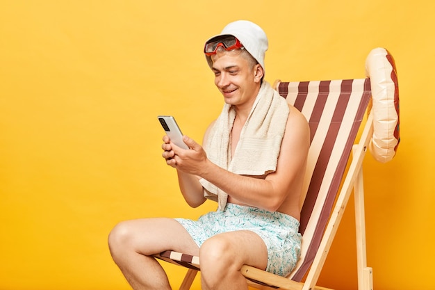 笑顔の幸せなシャツのない男がデッキチェアに座って黄色い背景で孤立し,ビーチリゾートで休んで,携帯電話を使ってソーシャルネットワークをチェックしています.