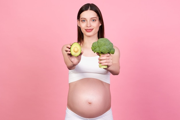 Smiling happy, slim, pretty white pregnant girl holding broccoli and avocado, posing with bare belly. Focus on vegetables. The concept of pregnancy, motherhood, preparation, expectation.