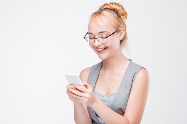 Smiling happy woman in grey vest and glasses typing message on mobile phone. Communication, chatting in social networks. Grey wall with copy space