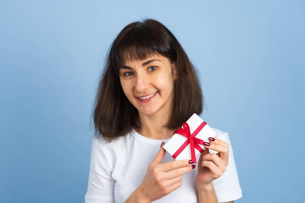 青い背景に分離されたギフトボックスを保持している幸せな女の笑顔。バレンタインデーや誕生日のコンセプト