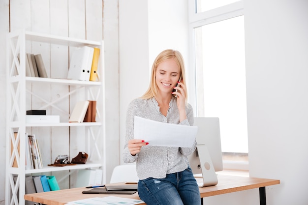 Smiling happy woman talking on mobile phone and holding document while sitting at her workplace