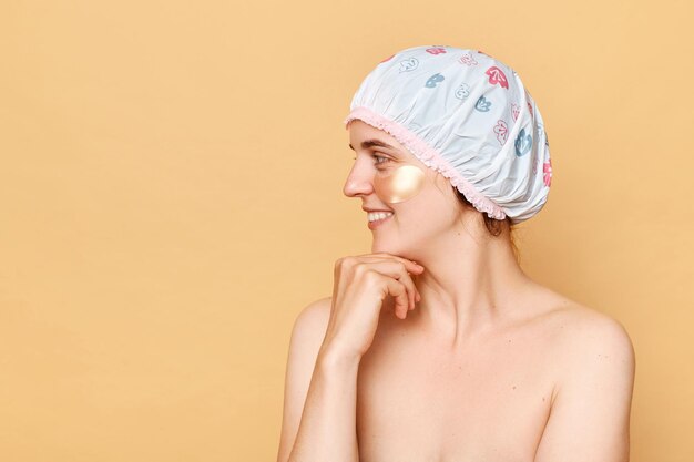 사진 홍보 텍스트를 위한 광고 공간을 위한 복사 공간을 바라보며 베이지색 배경 위에 격리된 눈 패치가 있는 샤워 모자를 쓰고 웃고 있는 행복한 여성