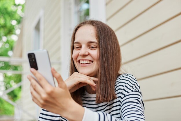 家の近くでポーズをとる電話を使ってストライプのシャツを着た笑顔の幸せな女性が、ビデオ通話をしながらスマートフォンで面白い動画を見ながらポジティブな感情を表現する