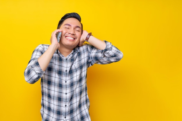 Smiling happy young Asian man wearing a white checkered shirt talking on a smartphone isolated on yellow background People Lifestyle Concept