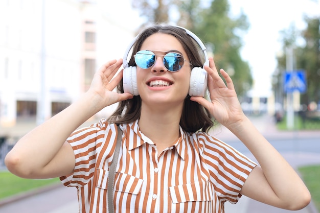 Smiling happy young beautiful woman in earphones walking outdoors in city.