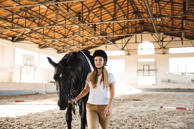 smiling happy young blonde beautiful woman wearing hat with horse in countryside in corral on training area.