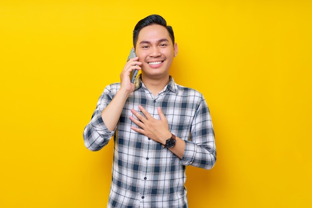Smiling happy young handsome Asian man wearing a white checkered shirt talking on mobile phone isolated over yellow background People Lifestyle Concept