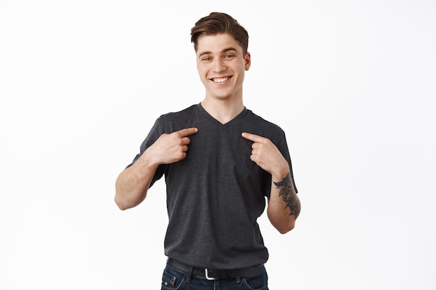 Smiling happy young man pointing at himself, nominating him, volunteer, choose pick me gesture, wants to participate, standing against white background